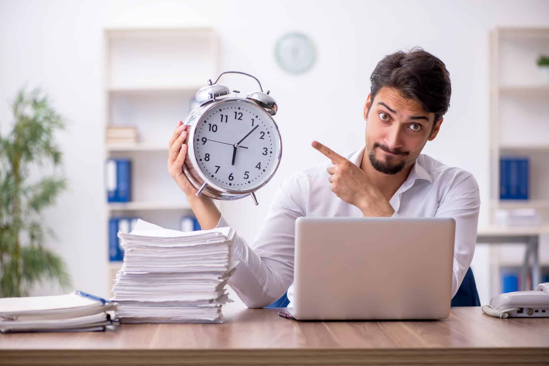 A man is sitting at his desk in front of his laptop with a large pile of paperwork. He is holding a large clock.