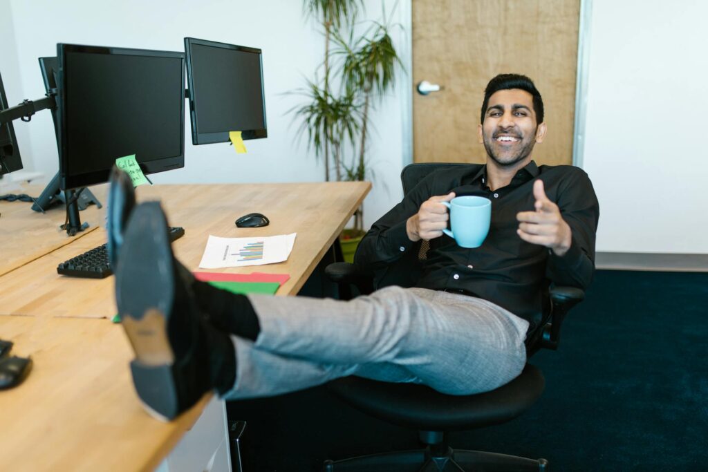 A man is leaning back in his office chair with his feet propped up on the desk. He is holding a cup of coffee.